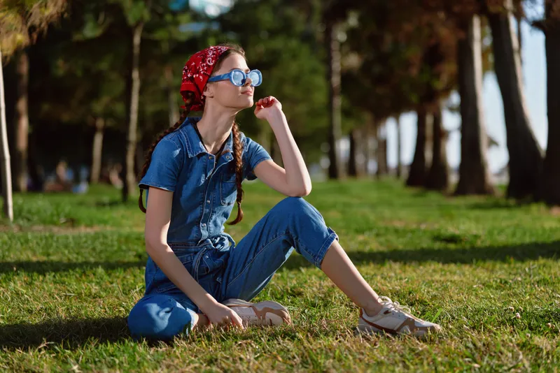 young woman sitting relaxed on grass wearing denim jumpsuit, red headscarf, sunglasses in sunny park with trees, casual outdoor style
