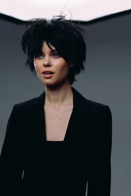 Young woman with curly black hair wearing a black blazer posing against a gray background