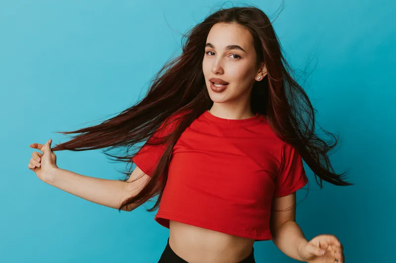 Young woman with long hair wearing red crop top posing against blue background, looking confident and playful with natural makeup and casual style in studio portrait photography.