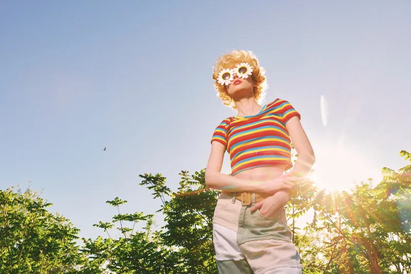 Smiling woman in a striped top outdoors under bright sunshine, showcasing a cheerful lifestyle with retro fashion and sunflower accents, captured in warm natural light.