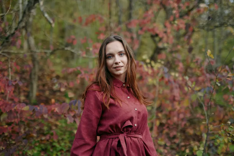 Candid portrait of a young woman in a burgundy coat standing in an autumn forest, captured with natural light and authentic detail, conveying realism and sincere presence.