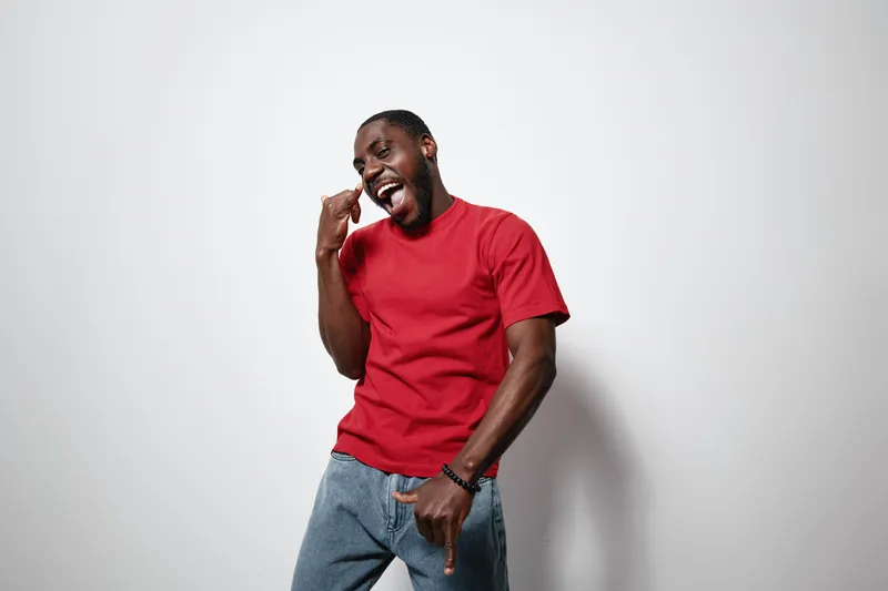African American man with dark skin wearing red t shirt and blue jeans posing with playful expression and hand gesture against white background in studio.