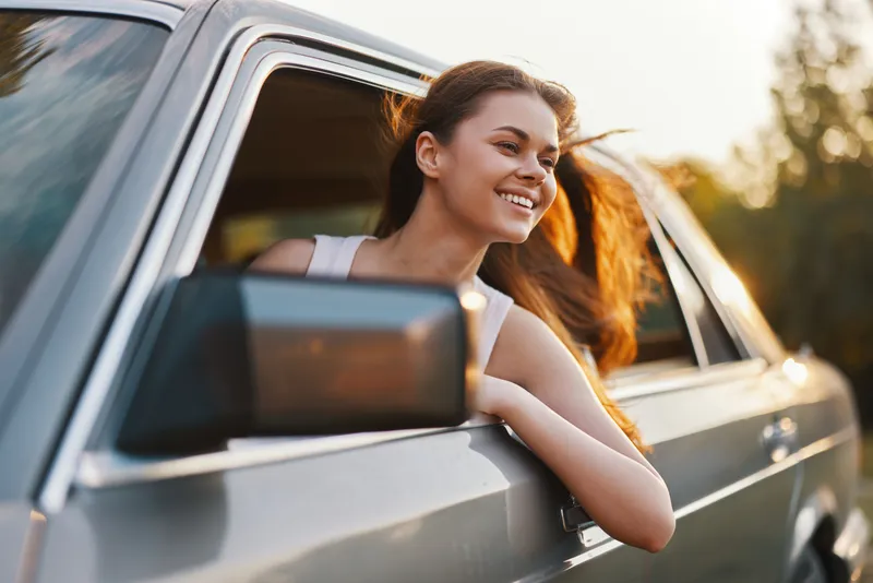 Cheerful young woman enjoying summer drive with wind in her hair, relaxed and happy, in a vintage car against a warm sunset backdrop.