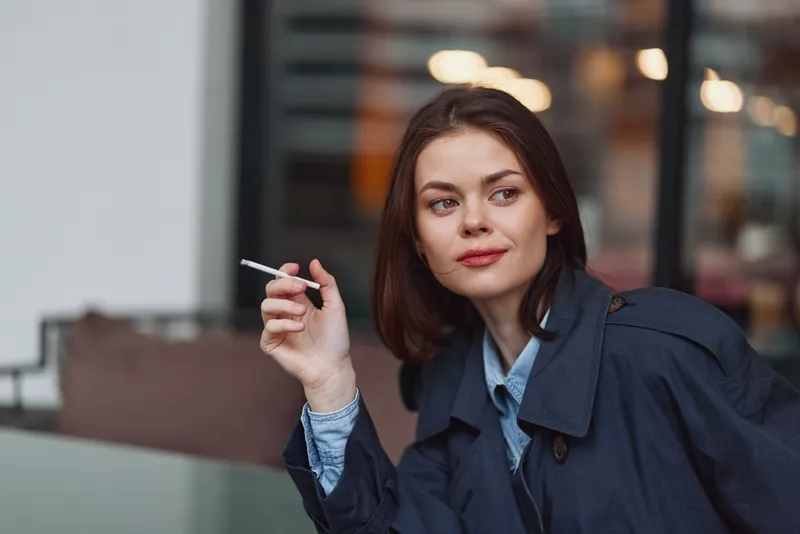 Young woman with brown hair wearing a dark coat holds a cigarette while sitting outdoors in an urban setting with soft evening lighting and a thoughtful expression.