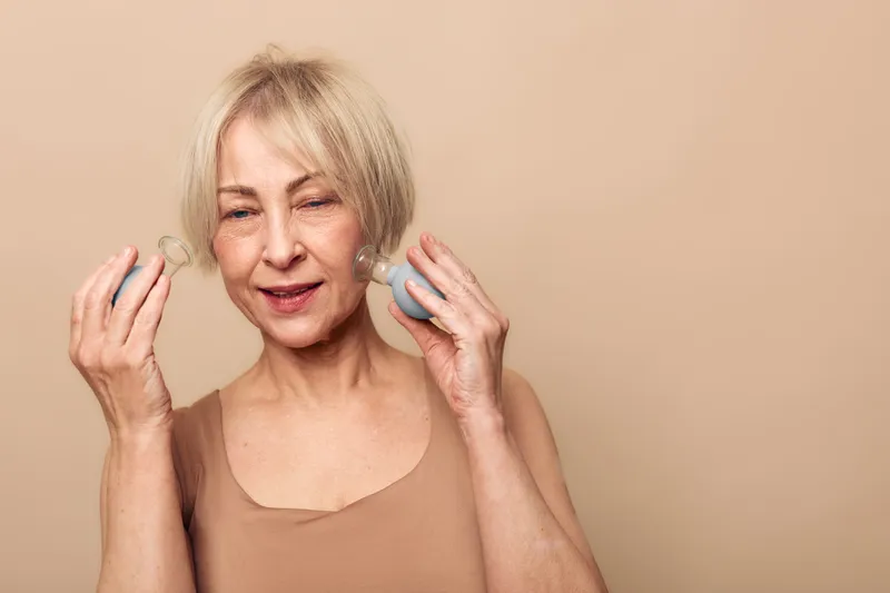 Mature woman with light hair uses facial massage tools on beige background. Skin care, anti aging, wellness concept with natural beauty and self care routine for healthy glowing skin.