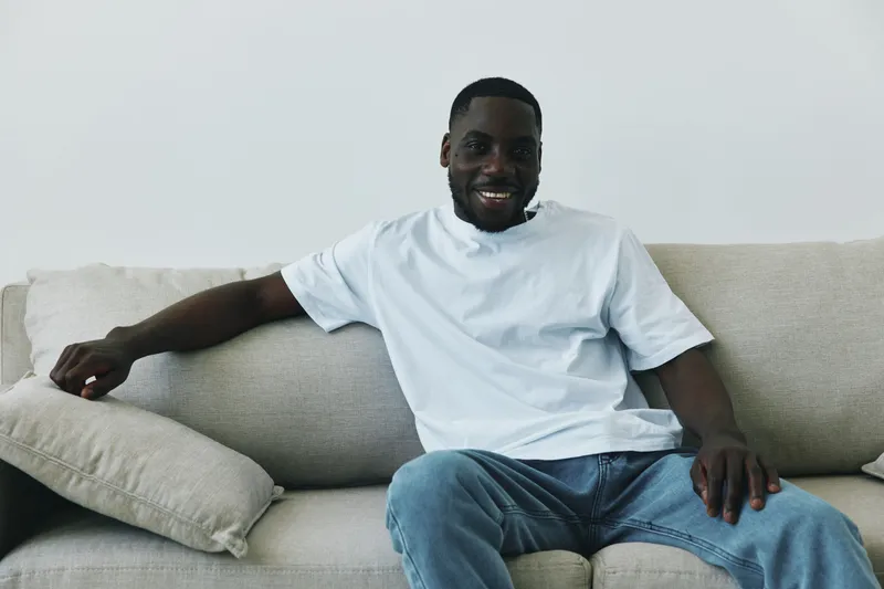 Happy young man smiling in casual white t shirt and jeans, sitting on a light sofa in a minimalist interior, embodying relaxation and comfort