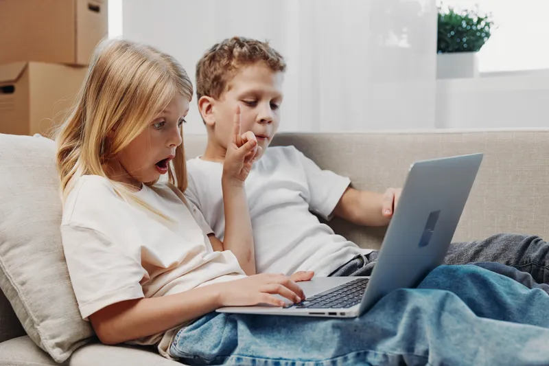 Two surprised children sitting on couch watching laptop screen, expressing shock and curiosity in bright modern living room environment.