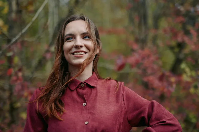 Authentic portrait of a cheerful woman in a burgundy shirt outdoors, natural light, candid smile, simple pose, genuine mood, clear focus, soft autumn backdrop.