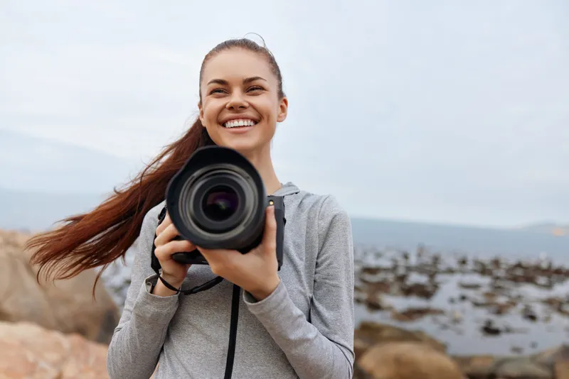 Happy female photographer with a camera on a rocky beach, showcasing joy and passion for photography during a cloudy day by the sea
