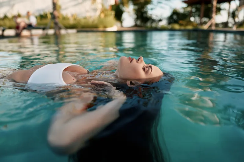 Carefree woman floating in a sunlit pool, eyes closed, relaxed silhouette, clear water reflections, summer leisure mood, outdoor calm, natural colors, tranquil moment as gentle breeze rustles nearby