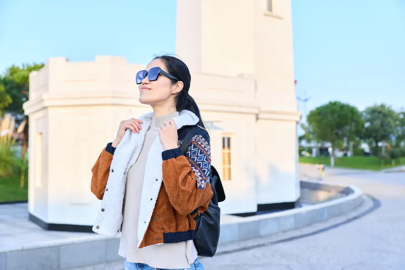Woman in sunglasses, scarf and brown jacket posing outdoors beside a monument, stylish travel style, bright day, urban scenery and confident mood in daylight