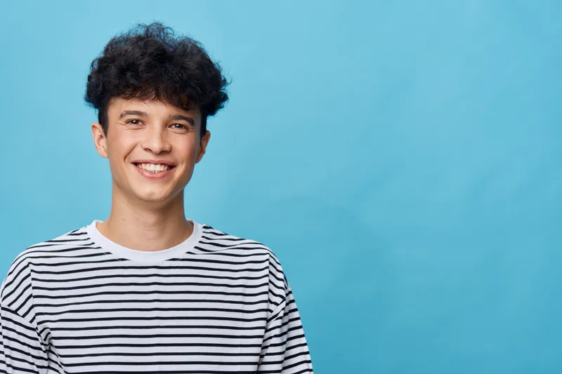 smiling man with curly hair wearing striped shirt posing against blue background, cheerful expression, casual style, young adult male portrait