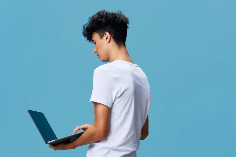 Young man using laptop with focus, standing against plain blue background, casual white t-shirt, technology and concentration concept, side view