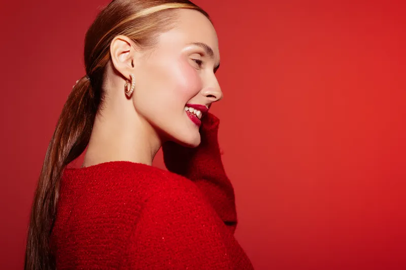 Young woman with long hair and red lipstick smiles wearing red sweater and gold earrings on red background. Concept of fashion, beauty, and confident female portrait.
