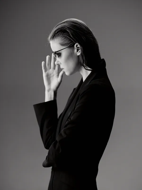 Black and white portrait of a thoughtful woman in glasses wearing a black suit posing sideways with hand raised