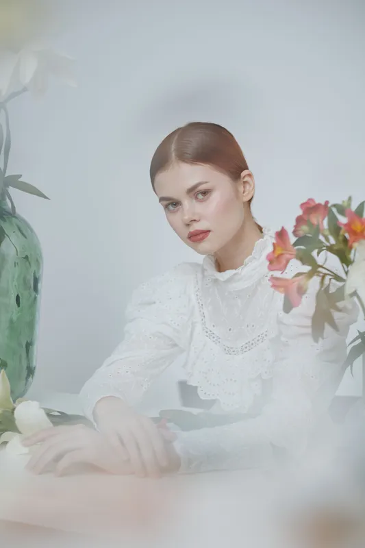 Elegant woman in white vintage dress with a serious expression, surrounded by soft floral arrangements, creating a serene and timeless atmosphere