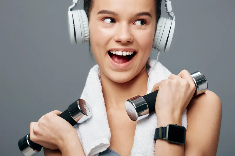 A fit woman lifts light dumbbells while wearing headphones and a towel draped over her shoulders, mid workout in a bright gym with focus on strength and energy.
