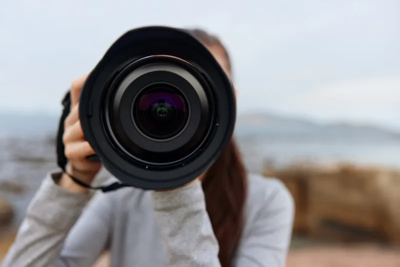 Young woman capturing nature with a camera, wearing casual gray clothing, conveying enthusiasm and creativity against a scenic outdoor backdrop