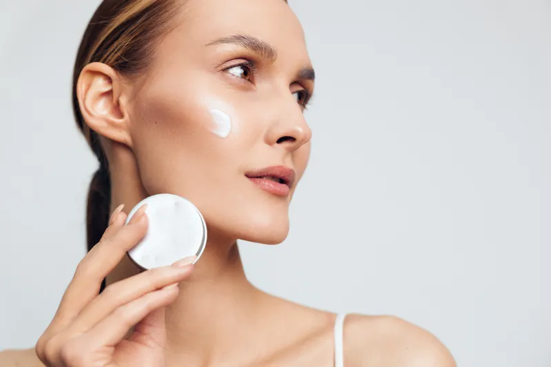 Young woman with clear skin applies white face cream on her cheek using a cotton pad against a plain light background, expressing calmness and skincare routine.