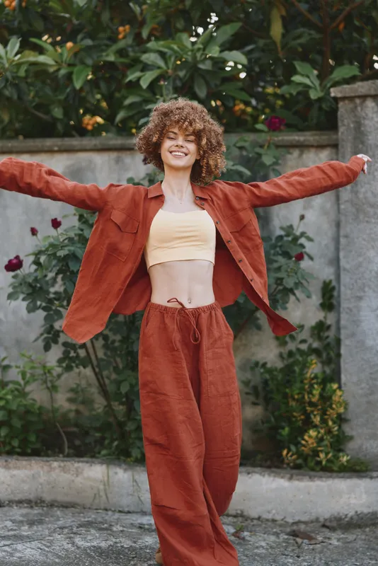 Cheerful woman outdoors, arms outstretched, sunlit lifestyle moment with a relaxed pose, warm colors, rust outfit, natural greenery backdrop, candid joyful mood and breezy atmosphere