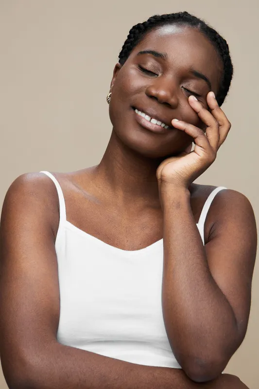 Beautiful young African woman in a white tank top, expressing joy and tranquility against a soft beige background, perfect for wellness and self care concepts
