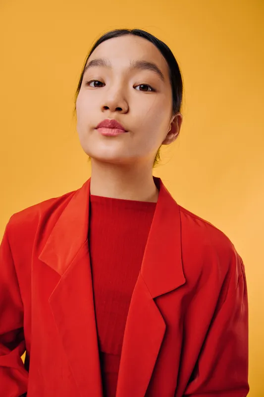 Portrait of confident woman in red jacket and sweater standing against yellow background. Young female model with natural makeup and black hair tied back wearing casual outfit indoors.