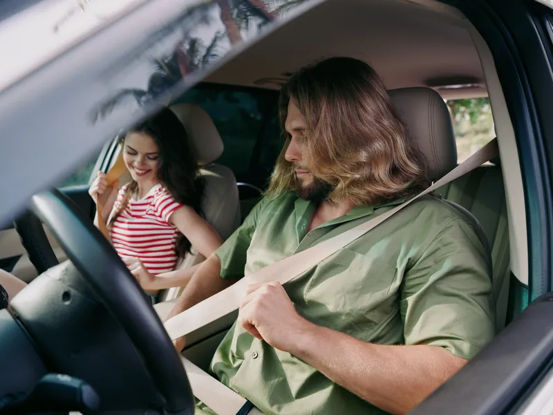 A man drives a car with a young woman in the passenger seat, both wearing seat belts. The scene emphasizes safety, focus, and everyday travel in a warm, natural interior.