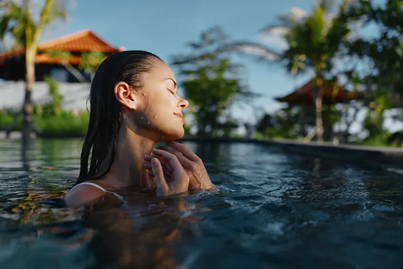 Relaxed poolside girl enjoys warm sunlight, serene water, tropical surroundings, and a gentle breeze, embodying leisure and calm in an outdoor resort.