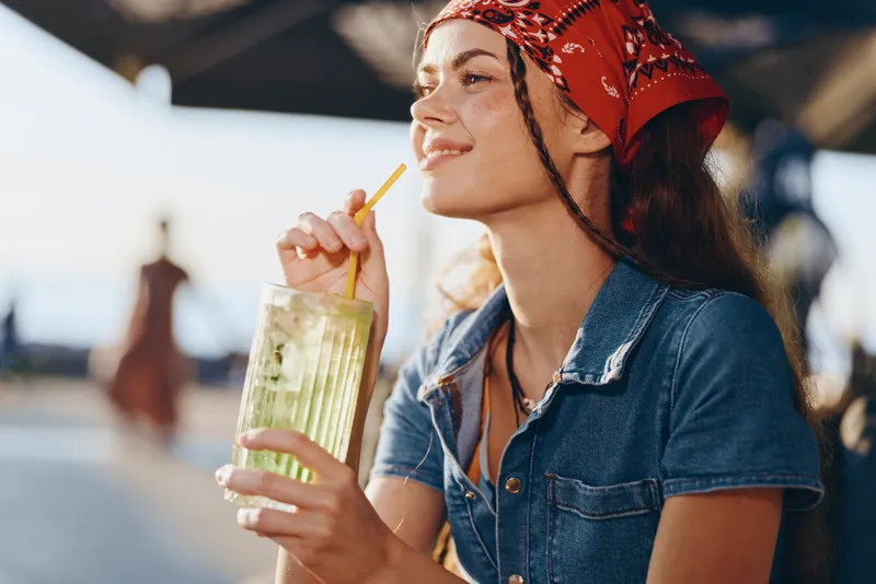 Lifestyle woman enjoying summer drink in modern boho-western denim street style, captured with warm film color tones and relaxed outdoor ambiance.