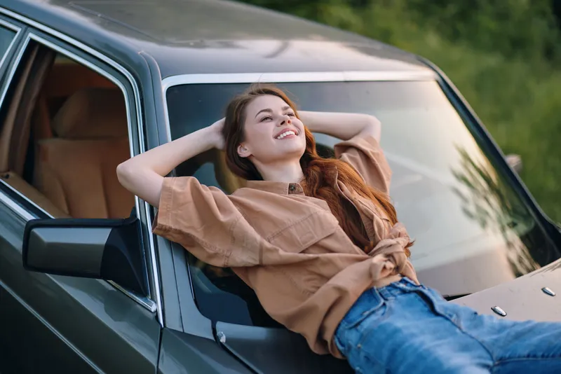 Young woman relaxing on the hood of a vintage car, smiling joyfully in natural light surrounded by green foliage. Casual attire, showcasing a carefree summer vibe.
