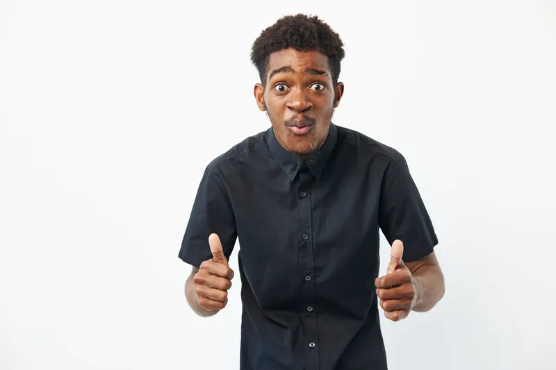 Black man showing enthusiasm with thumbs up, wearing a black shirt, expressing joy in a simple, bright environment that enhances the positive vibe.