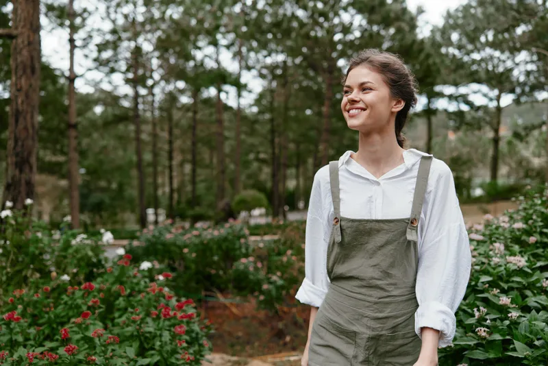 Joyful young woman in green overalls walking through a blooming garden, surrounded by vibrant flowers and lush trees, reflecting a serene connection with nature