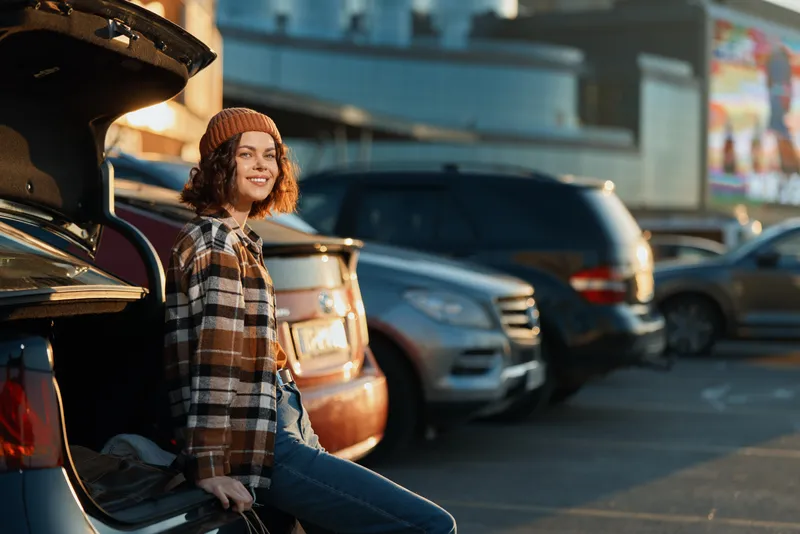 woman sitting on car trunk in parking lot, smiling with golden urban light, candid authenticity and mindful living vibe, cozy beanie and plaid jacket capturing emotional storytelling moment
