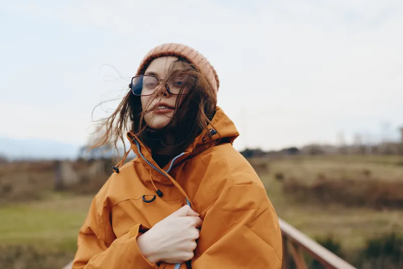Woman lifestyle portrait outdoors in winter, wearing an orange jacket, knit hat and glasses, standing on a rural path, wind moves hair, hand at collar, soft daylight fields