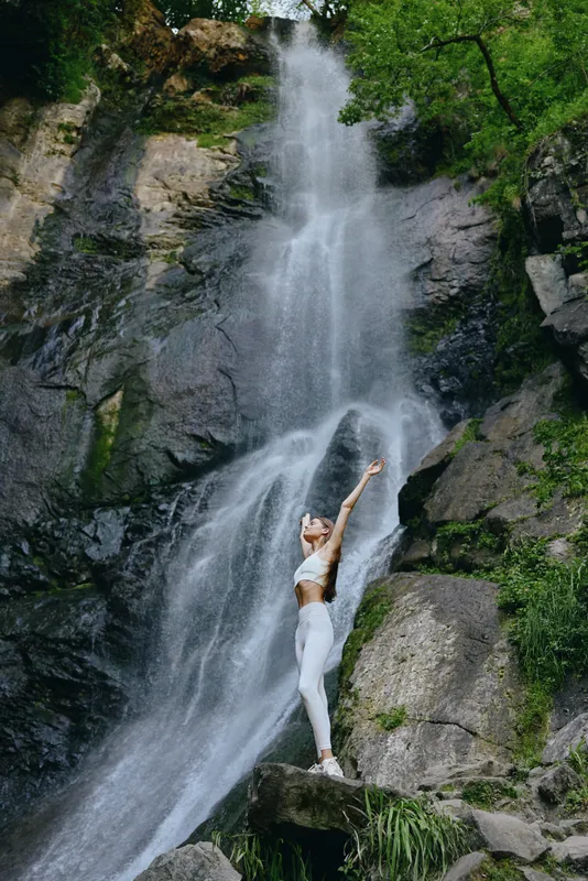 woman stretching near waterfall, outdoor fitness activity, natural green environment, healthy lifestyle, athletic yoga pose, serene mountain setting