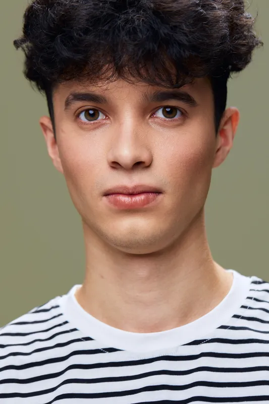 Young man with curly hair wearing striped shirt looks directly at camera with neutral expression, soft lighting highlights his facial features and warm skin tone.