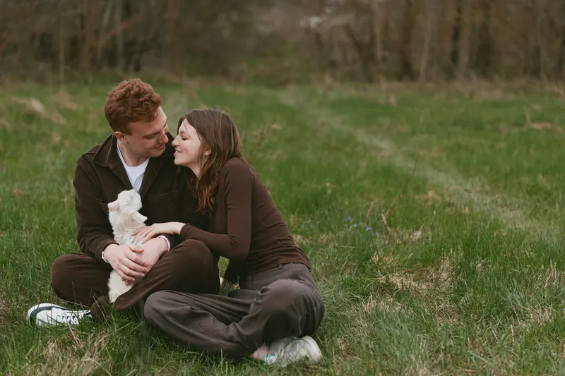 A couple sits on grass in a sunny field, sharing affection with a fluffy puppy, smiling and relaxed, capturing a casual lifestyle moment outdoors, warm light, candid romance, simple joy