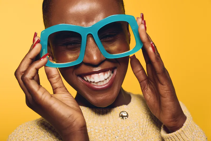 Smiling dark skinned African woman wearing large blue glasses and yellow sweater enjoys bright studio portrait on yellow background. Concept for joy, fashion, and positive emotions.