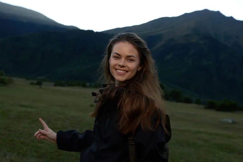 A cheerful young woman stands in an open field with distant mountains, smiling at the camera and radiating confidence against a natural, scenic backdrop.