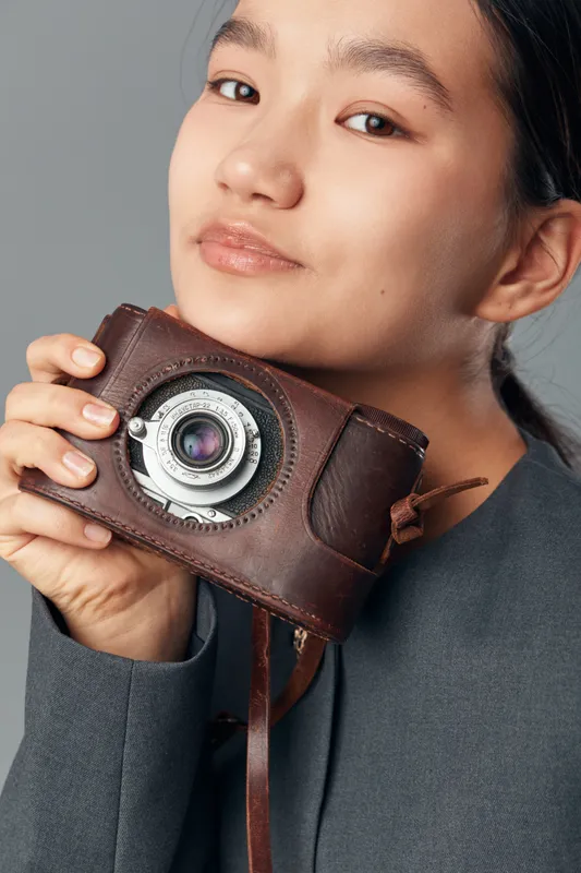 Young woman holding vintage camera with leather case close to her face, studio portrait with neutral light and grey background. Female photographer, classic equipment, retro style.