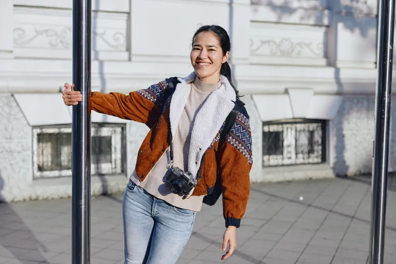 Woman outdoor fashion casual jacket scarf smile on street with urban architecture, posing beside pole, candid street style, bag accessory, daylight and breeze in modern city vibe