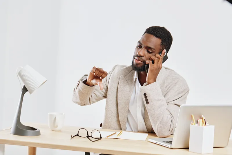 Businessman making a phone call at his modern desk with laptop, notebook, mug, and glasses, cheerful and professional, indoor office setting, casual dress, light background, workspace communication co