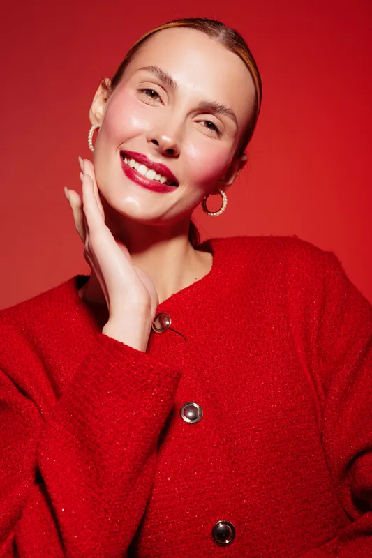 Smiling woman with red lipstick and hoop earrings poses in red sweater against red background. Close-up portrait with natural makeup and joyful expression. Concept of beauty and fashion.