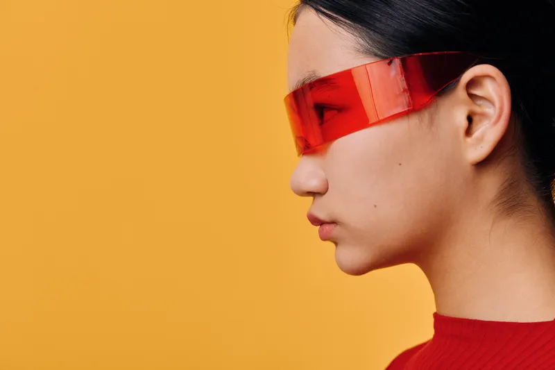 Profile of young woman wearing futuristic red visor and red top against solid orange background. Female with sleek hair and modern accessory in studio fashion portrait.