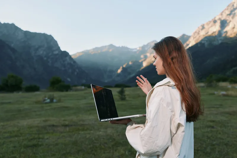 A focused professional woman stands outdoors with a laptop in a scenic mountain valley. She looks at the screen with calm concentration, creating a productive and tranquil remote work atmosphere.