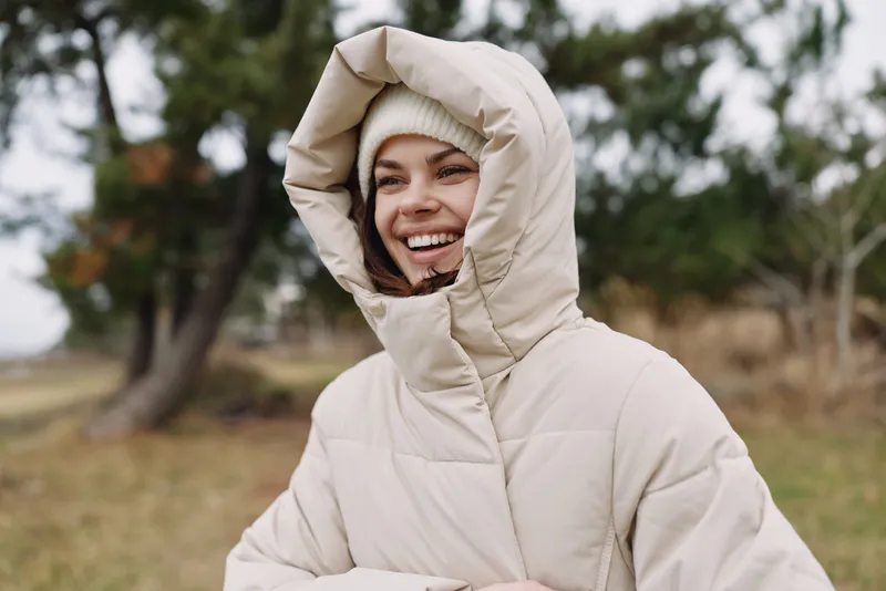 Woman outdoors wearing a beige puffer coat with hood up, smiling and enjoying crisp weather in a park setting, her joyful expression captures warmth against a chilly day with soft natural light,