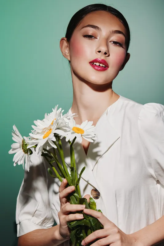 Fashionable young woman with glossy lips and natural makeup holds fresh daisies wearing an elegant white blouse. Studio shot with soft light and pastel wall background.