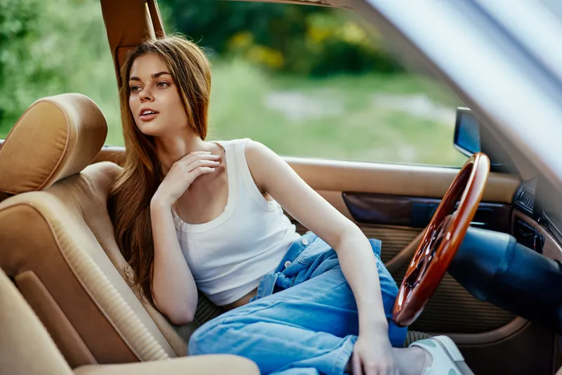 Young woman in casual clothing sitting in a vintage car, enjoying nature, with a relaxed expression, conveying a sense of freedom and tranquility during summer days.