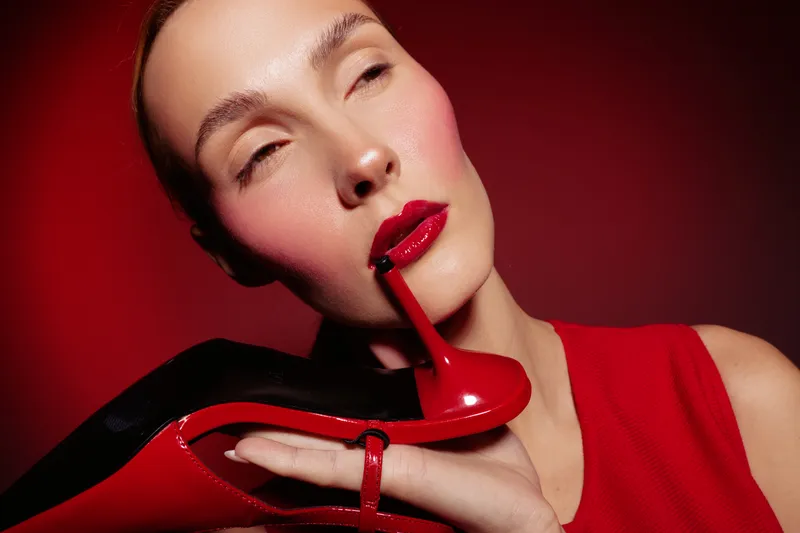 Close-up of a woman with red lipstick holding a red high heel shoe near her face against a red background. Concept of fashion, beauty, and bold style with vibrant colors.