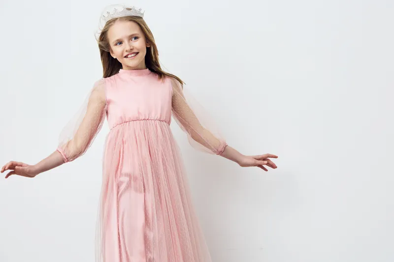 Young girl in a pink tulle dress, wearing a crown, joyfully posing against a white background, embodying innocence and elegance, perfect for childhood celebrations